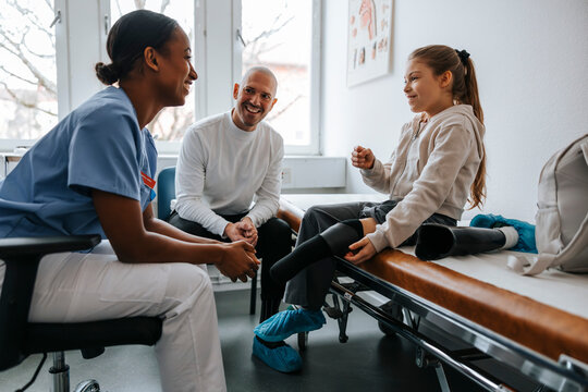 Smiling girl with artificial limb talking to female medical professional while sitting on bed in examination room