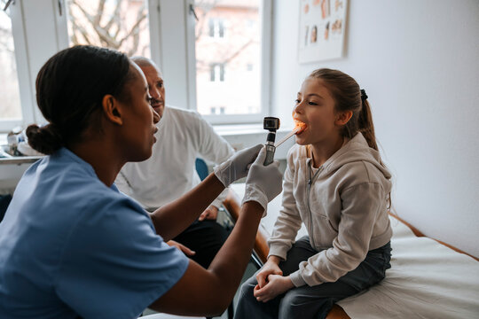 Female pediatrician examining throat of girl using tongue depressor and flashlight in examination room