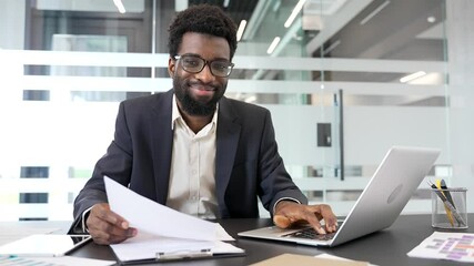 Portrait of a happy African American businessman sitting at workplace in business office. Head shot of smiling financier in formal suit working at a desk with a laptop and papers and looking at camera