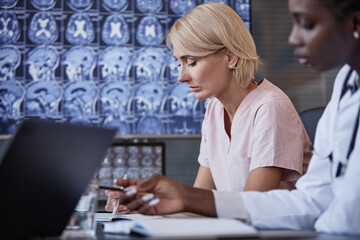 Female radiologist writing notes in notebook thoroughly studying anamnesis of patients disease in discussion with colleagues at meeting table with brain CT images on screen in backdrop, copy space