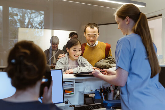 Female medical worker booking appointment for father and daughter behind glass partition at hospital