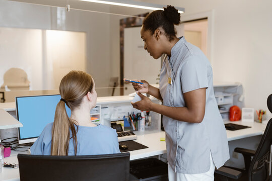 Female nurse discussing with receptionist colleague sitting at desk in hospital
