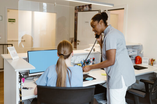 Side view of female healthcare professional attending call while standing next to receptionist at hospital