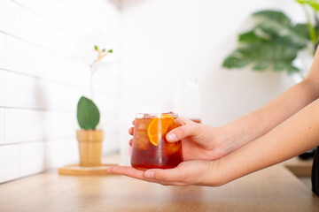 Woman's hands holding a cup of fresh cold brew coffee. The glass is filled with ice cubes with and slice of orange. Wooden kitchen counter with white tiles and green plants in backgroung.