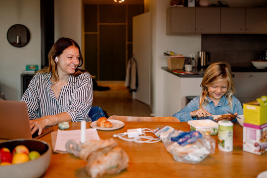 Smiling mother with laptop watching son spreading butter on bread at home