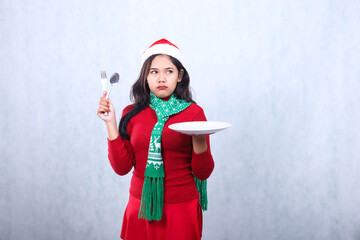 expression woman wearing red sweater red sweater with santa hat and scarf celebrating merry christmas, annoyed to the left carrying white plate and holding spoon fork, isolated on white background