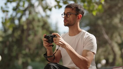 A young man with glasses sits on a bench in a park reviewing photos he took with a vintage film camera. He is wearing a white t-shirt and dark pants.