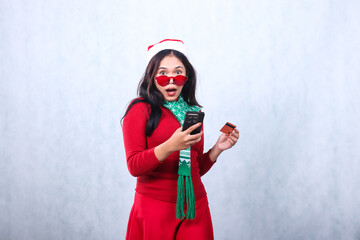 young asian woman surprised looking at camera holding gadget screen, credit card, wearing glasses, wearing red christmas sweater with santa hat and scarf, isolated on white background