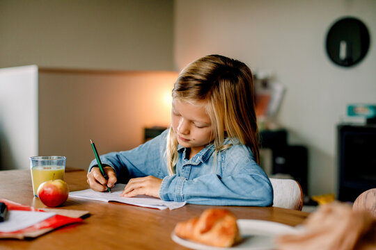Focused boy with long hair writing on book while dong homework at home