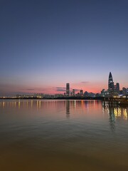 Obraz premium A serene twilight scene along the riverbank , with city lights beginning to twinkle in the distance and silhouettes of tall buildings against the backdrop of a clear sky.