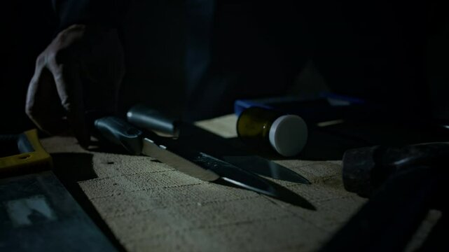 Closeup of hand of unrecognizable killer choosing murder weapon in dark room, taking sharp knife from table and checking it