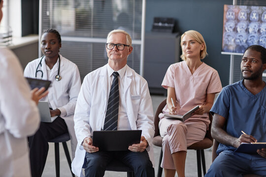 Senior male head physician listening to colleagues presentation with diverse team of doctors while participating in seminar at conference room