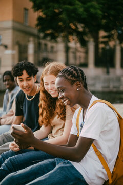 Happy teenage boy with braided hair showing smart phone to female and male friend