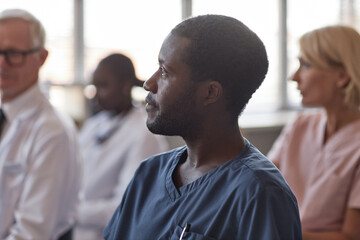 Side view portrait of African American man in blue scrubs engaged in colleagues presentation at doctors conference in meeting room while taking continuing learning course at hospital, copy space