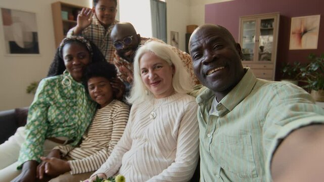 UGC shot of extended family of six members smiling and waving at camera while relaxing on couch in living room