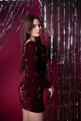 A confident woman dressed in a sparkling maroon outfit poses elegantly behind shiny silver curtains. The creative backdrop enhances the vibrant atmosphere of the indoor event.