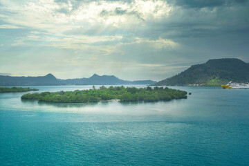 A body of teal water with islands, lush green vegetation, and a ship in the distance. Cloudy sky.