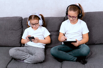 Two little sisters doing homework with a smartphone, sitting on the couch at home. A convenient and modern way to learn in a comfortable environment.