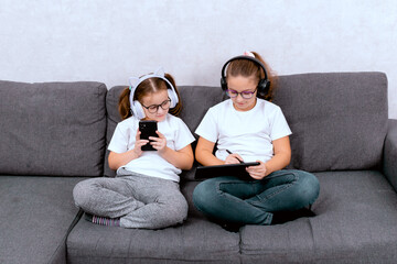 Two little sisters doing homework with a smartphone, sitting on the couch at home. A convenient and modern way to learn in a comfortable environment.