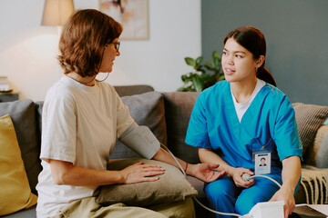 Fototapeta premium Nurse wearing blue scrubs and ID badge taking blood pressure of senior patient on couch using blood pressure cuff in living room with plant background