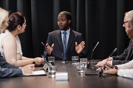 Medium shot of confident African American male presidential candidate speaking with political party committee developing campaign strategy while sitting at table against black curtains, copy space