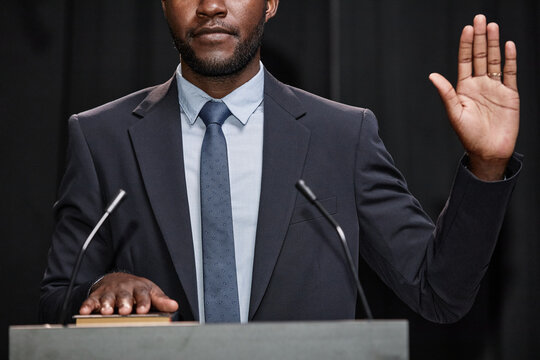 Cropped shot of African American male politician in blue suit standing at podium with one hand on book and another raised in oath to abide by law on stage, against black curtains - Powered by Adobe