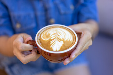Hot coffee mug, latte art on coffee, leaf-shaped milk foam, beautiful hot latte art in woman's hands. Top view of coffee art on wooden table from professional barista artist.