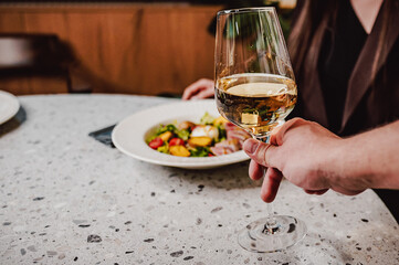 A close-up of a hand holding a glass of white wine, with a blurred background featuring a person and a beautifully arranged salad on a marble table. Perfect for food and dining themes.