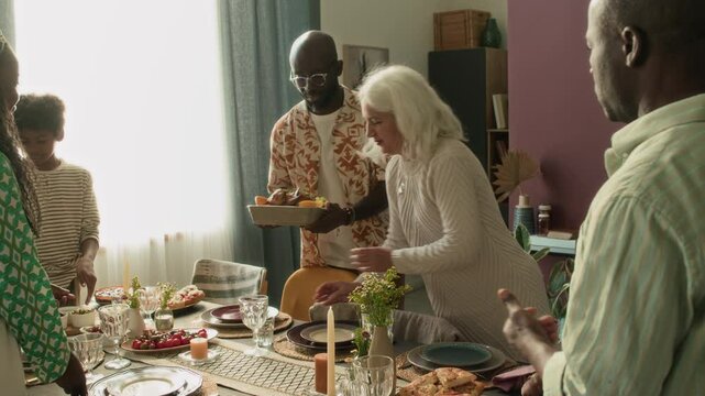 Tilt up shot of multigenerational family arranging table for holiday lunch, young man bringing baked chicken and putting dish in center