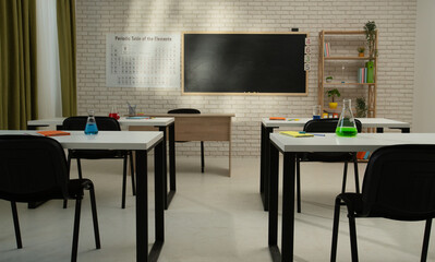 Empty school chemistry classroom with desks, chairs and chalkboard, workspace for teaching and studying. Glass bottles with liquids on tables, back view.
