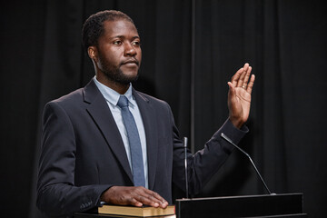 African American male politician dressed in blue suit keeping one hand on book and another raised swearing to obey law while standing at podium on stage against black curtains, copy space