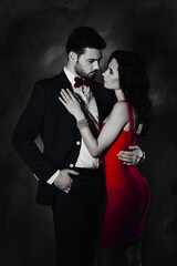 Portrait of bearded man in tuxedo with bow holding hand in pocket of pants and charming pretty lady with booty ass jewelry, attractive spouses hugging looking at each other over grey background