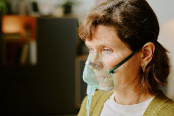 Portrait of an elderly woman wearing an oxygen mask while sitting indoors and looking straight ahead, showing her need for respiratory support and the clinical care provided