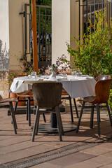 A round table is elegantly set with dishes, surrounded by wooden chairs on a sunny day, complemented by lush greenery in the background