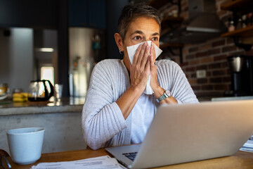 Senior woman sneezing while working on laptop at home