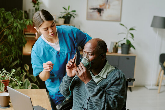 Healthcare professional assisting black elderly man with oxygen mask, scene showing interaction and care in well-lit room with plants and medical equipment