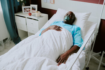 Elderly Black person lying in hospital bed, wearing oxygen mask and blue hospital gown, with...