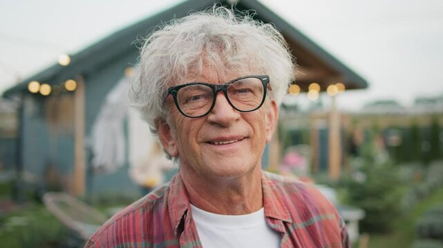 Close facial view of Caucasian male with glasses and grey hair. Man looking to side and glancing at camera while smiling. Wearing red checkered shirt. Standing in garden next to small house.