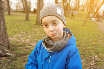 A young boy wearing a warm jacket and hat, gazing pensively in a park setting. Captures the charm of childhood, emotions, and the beauty of an autumn or early spring day outdoors