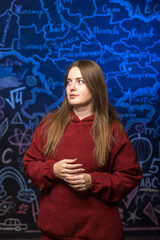 A woman in a red sweatshirt poses against a chalkboard filled with a map, equations, and scientific...