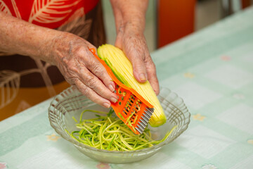 Close up nas mãos de uma cozinheira ralando uma abobrinha para uma tigela sobre a mesa. © Angela