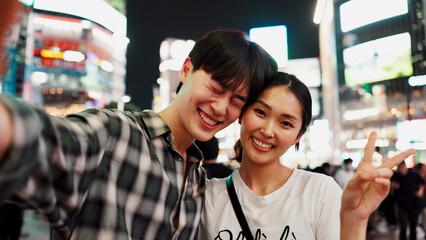 City, peace sign and selfie at night with couple in Japan together for bonding, love or tourism. Emoji, photograph and smile of happy Japanese woman with man on street for holiday, travel or vacation