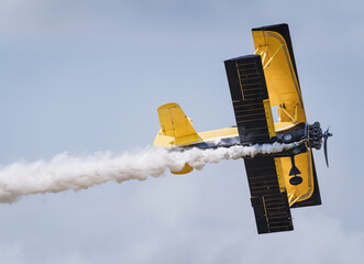 A close-up view from below of a yellow biplane performing maneuvers in the sky at an air show, leaving a smoke trail behind. © Skatty