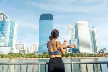 Back of Asian sport woman do arm stretching near wall railing and look forward to relax after exercise in public garden with high building of big city in background and soft light.