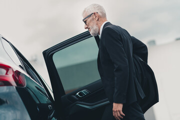 Elderly businessman in formal suit getting into car on city street, symbolizing travel and professionalism