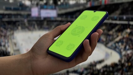 GREEN SCREEN CHROMA KEY A hand holding a phone with a green screen at a hockey game in an indoor stadium, ideal for showcasing live sports or betting apps