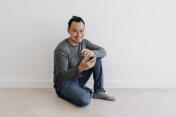 Happy creative freelance Asian man sitting on the floor using his phone.