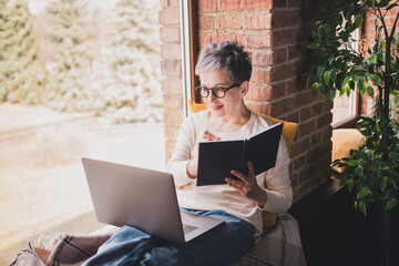 Elderly woman with grey hair sits by window reading book and using laptop in cozy home interior during the day