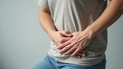 A person in a gray t-shirt is sitting down and holding their stomach, indicating discomfort or pain. The background is plain and neutral, reflecting a sense of unease and distress