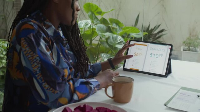 African American female marketer comparing conditions for subscription services zooming them in on digital tablet while drinking coffee at work desk